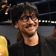 A middle-aged Japanese man in thick, circular glasses smiles at something off-camera.